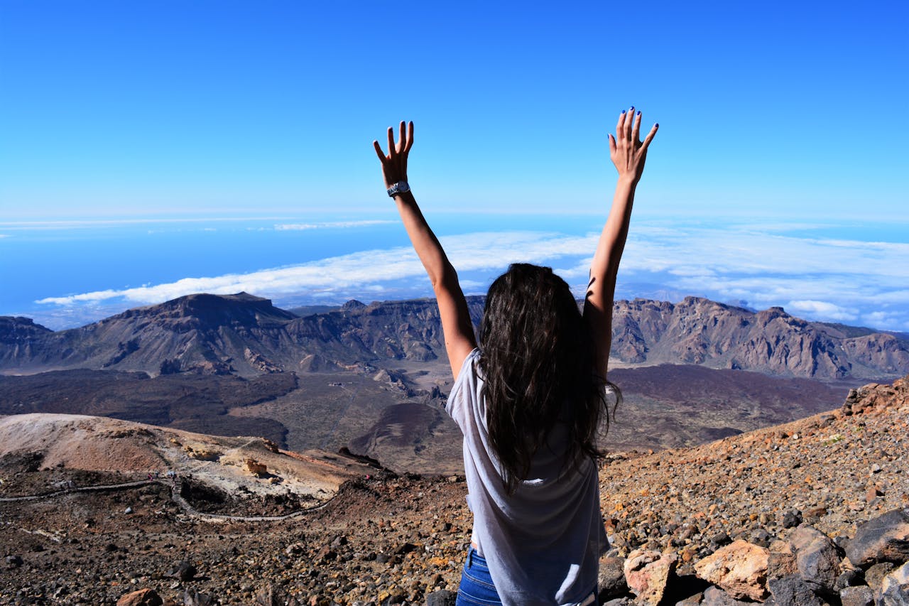 A woman celebrates atop Mount Teide with arms raised, overlooking the vast landscape and sea.