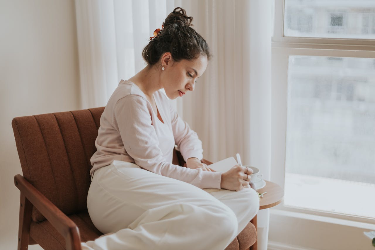 hero-img-02 A woman comfortably journaling by a window, surrounded by soft natural light, in a cozy indoor setting.